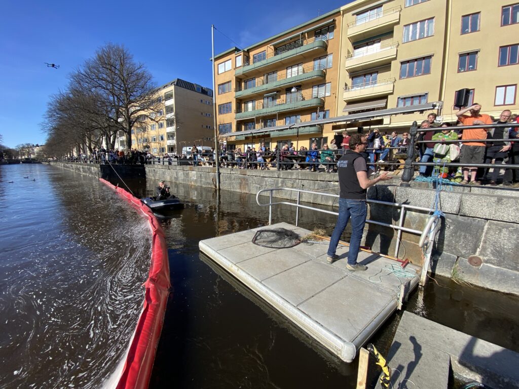 En man står på en flytbrygga i ett vattendrag i stan framför en stor grupp människor.