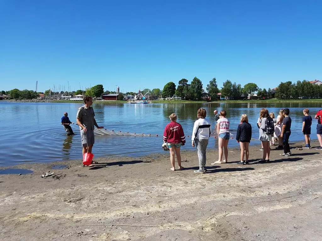 Flera barn står på en sandstrand, i vattnet utanför står två personer med vadarbyxor.