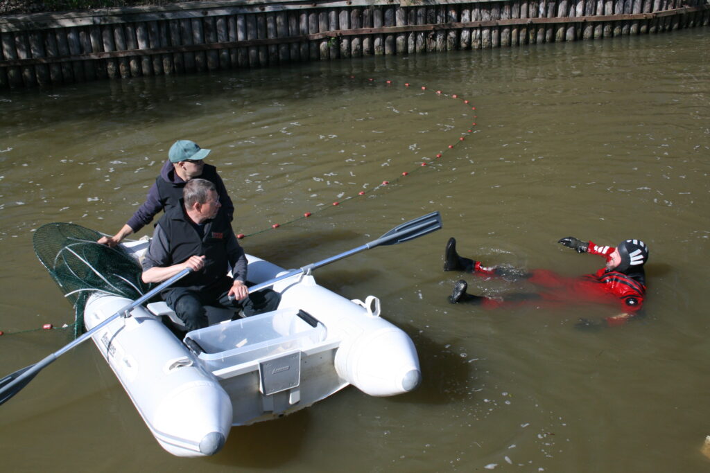 Två personer sitter i en gummibåt på en å, en person med vådträckt ligger i vattnet.