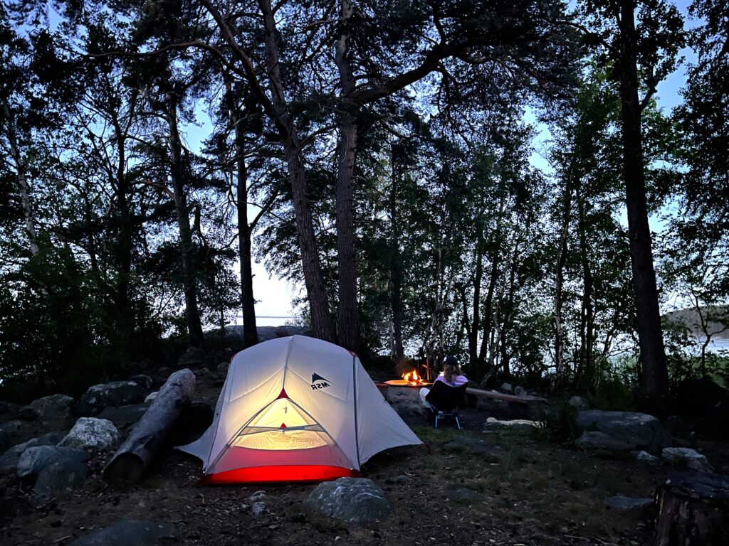 Ett tält uppsatt i en mörk skog. En lampa i tältet gör att tältet lyser.
